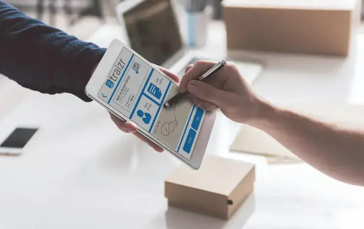 Person signing for a parcel using the Traizr app on a smartphone, with cardboard boxes in the background showing workplace package delivery.